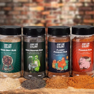 May include: Four spice rub jars with black lids and labels featuring cartoon illustrations. The jars are labeled "Good Boy Rub," "Grim Reaper Rub," "Flabby Phoenix Rub," and "Pigasus Rub." Each jar contains a different colored spice blend, with the text "How Low Slow?" on each label.