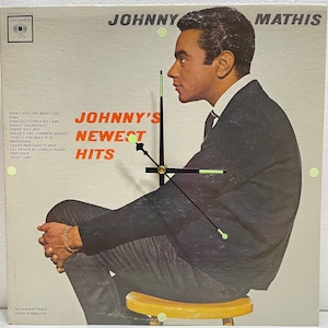May include: A vintage vinyl record album cover featuring a black and white photograph of Johnny Mathis, a popular American singer. The album title is "Johnny's Newest Hits" and lists the songs included on the record. The album cover is a light grey colour with a black and white photograph of Johnny Mathis sitting on a chair. The album cover is a popular collectible for music lovers.