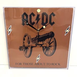 Könnte beinhalten: Quadratische Wanduhr mit braunem Hintergrund mit dem AC/DC-Logo und einer Kanonenillustration. Die Uhr hat schwarze Zeiger und weiße Blitzakzente. Der Text "FOR THOSE ABOUT TO ROCK" steht unten.