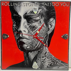 May include: A red vinyl record with a black and white image of a person with a tattoo on their face. The record has the text "Rolling Stones Tattoo You" on the top and four guitar picks attached to the corners.