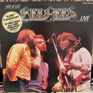 May include: Album cover for "Here at Last... Bee Gees... Live." The cover features a photo of the Bee Gees performing live. The album title and band name are in a stylized font. A sticker indicates the album includes 22 Bee Gees all-time greatest hits.