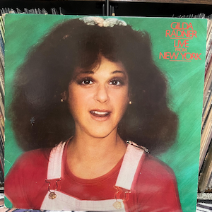 May include: Album cover featuring Gilda Radner with the text "Gilda Radner Live From New York" on a green background. Radner is wearing a red shirt and white overalls. Her hair is dark and voluminous.
