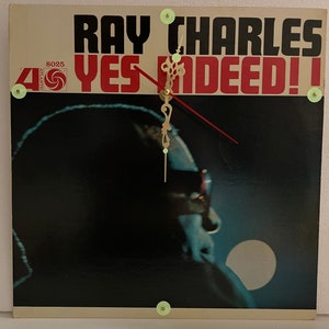 May include: A vintage vinyl record clock featuring a black and white photo of Ray Charles. The clock face is white with black hands and the record label is visible with the title "Yes Indeed!" and the number 8025.