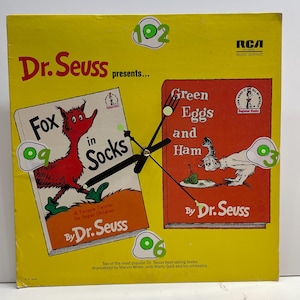 May include: A yellow Dr. Seuss clock featuring the covers of "Fox in Socks" and "Green Eggs and Ham." The clock has black hands and egg-shaped hour markers. The clock is a vintage RCA Music Service product.