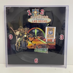 May include: Square wall clock with a black background featuring a Las Vegas theme. The clock face includes a motorcycle graphic, a Frank Sinatra poster, and the words "Welcome to Fabulous Las Vegas."