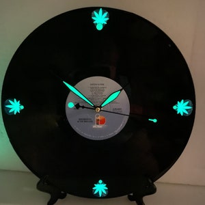 Peut inclure: Une horloge en vinyle noir avec des accents en forme de feuille de marijuana qui brillent dans le noir. Le disque est "Catch a Fire" de Bob Marley & The Wailers. Les aiguilles de l'horloge sont noires et dorées.