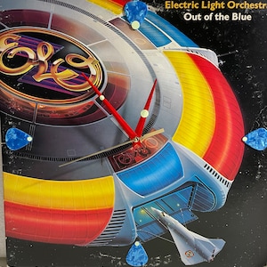 May include: A clock made from a vintage Electric Light Orchestra album cover, featuring the album title "Out of the Blue." The clock has red hands and is decorated with blue guitar picks. The album art depicts a spaceship design with a retro aesthetic.
