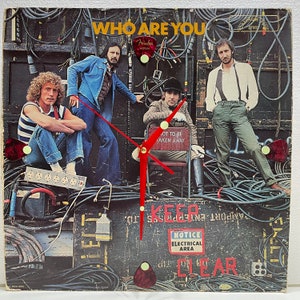 May include: A vintage vinyl record album cover featuring the band 'Who' with the album title 'Who Are You' in white text on a black background. The band members are standing in front of a black backdrop with electrical cords and equipment. The album cover has a red clock hand with a gold center and a red clock face with the words 'Keep Clear' in white text.