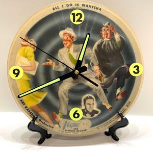 May include: A vintage-style clock featuring a retro illustration of people dancing. The clock face has a black background with the numbers 3, 6, 9, and 12 in yellow. The clock hands are black with neon green tips. The clock is mounted on a black stand.
