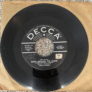May include: A black vinyl 45 RPM record by Bill Haley and His Comets featuring the song Rock Around the Clock on a Decca label. The record has a central circular hole and a paper label with white text, including the record number 9-29124.