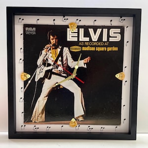 May include: A square wall clock featuring an Elvis Presley Madison Square Garden album cover as the face. The clock has a black frame, gold-coloured guitar pick hour markers, and gold hands with a red second hand. Musical notes border the white edge.
