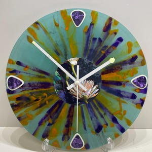 May include: A unique wall clock made from a repurposed vinyl record. The clock face features a teal, orange, and purple design with a floral centerpiece. White clock hands and purple accents complete the artistic look. The clock is displayed on a clear stand.