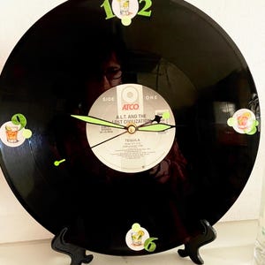 May include: A black vinyl record clock with green clock hands and numbers. The clock face features the label of the record "Tequila" by A.L.T. and the Lost Civilization. Decorative cocktail stickers are placed around the clock face.