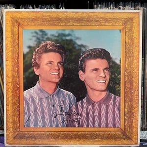 May include: Vintage vinyl record album cover featuring a framed portrait of two men in patterned shirts. The album cover has a gold-colored frame and includes handwritten signatures. The background shows a blue sky and green foliage.