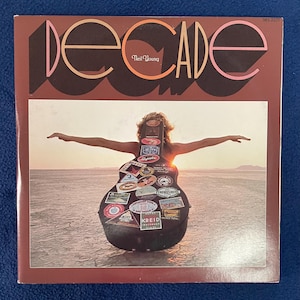 Könnte beinhalten: Albumcover für "Decade" von Neil Young. Das Cover zeigt das Wort "Decade" in großen, stilisierten Buchstaben. Darunter ist eine Person mit ausgestreckten Armen vor einem Sonnenuntergang zu sehen, mit einem gitarrenförmigen Objekt, das mit Aufklebern bedeckt ist.