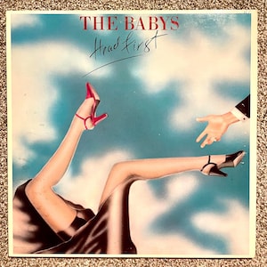 May include: A square vinyl record cover titled The Babys Head First featuring an illustration of legs in a black skirt against a blue sky. One foot wears a red high heel, the other a black high heel. A hand reaches toward the legs from the right.