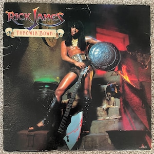 May include: A vintage vinyl record cover for Rick James Throwin' Down featuring a person with long dark hair holding a bass guitar shaped like an axe, wearing a gold necklace, a wide belt, and black leg guards while holding a round metal shield.