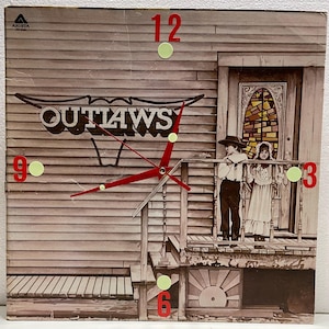 May include: A vintage vinyl record album cover featuring a red and white clock face with the word "OUTLAWS" in a stylized font. The clock is set against a backdrop of a wooden house with a porch and a stained glass window. Two children stand on the porch, a boy in a black hat and a girl in a white dress.