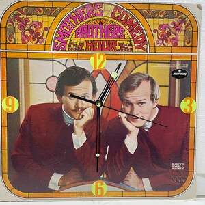 May include: A vintage vinyl record album cover featuring a clock face with the words "Smothers Brothers Comedy Hour" and a photo of the two brothers. The album is by Mercury Records and Rubicon River Records.