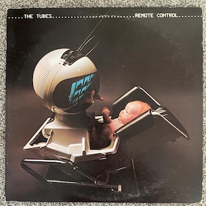 May include: A square vinyl record cover featuring the text THE TUBES REMOTE CONTROL at the top. The image shows a white, spherical, vintage-style television set integrated into a black and silver infant seat, with a baby sitting inside.