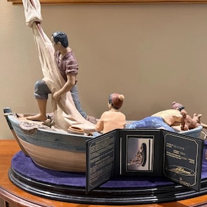 Può includere: Statuetta nautica dettagliata raffigurante una barca con tre figure. Una figura tira una vela, un'altra è seduta e una terza è sdraiata. La barca è su una base rotonda con una copertura in velluto viola. Include un'immagine incorniciata.