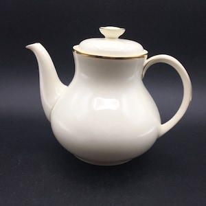 Puede incluir: Tetera de cerámica color crema con cuerpo redondeado, boquilla curva y asa de bucle. La tapa está rematada con un pequeño pomo. Los adornos dorados acentúan el borde y el asa. La tetera está sobre un fondo oscuro.