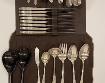 Op de afbeelding: Een set antiek zilverwerk, waaronder vorken, lepels en messen, gerangschikt op een donkerbruine stof. Het zilverwerk heeft sierlijke handvatten en een glanzend oppervlak. De set bevat opscheplepels en botermessen.