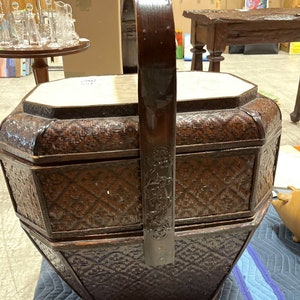 May include: A dark brown, octagonal, woven basket with a tall, arched handle. The basket has a decorative, carved handle and a light-colored top. The basket is sitting on a blue surface.