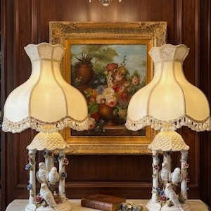 May include: Two ornate table lamps with cream-colored shades and decorative bases featuring bird figurines. The lamps sit on a marble-topped table in front of a framed painting and a dark wood panel wall. A crystal chandelier hangs above.