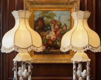 Puede incluir: Dos lámparas de mesa ornamentadas con pantallas color crema y bases decorativas con figuras de pájaros. Las lámparas están sobre una mesa con tapa de mármol frente a una pintura enmarcada y una pared de paneles de madera oscura. Un candelabro de cristal cuelga encima.