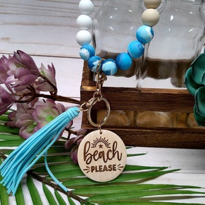 May include: A beaded keychain wristlet with a light blue tassel and a wooden charm. The charm is engraved with the words "beach please" and a sunburst design. The beads are white, tan, and blue.