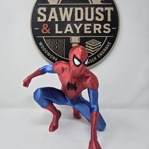 Puede incluir: Una figura de Spider-Man impresa en 3D en pose agachada con un traje rojo y azul met&aacute;lico y un emblema de ara&ntilde;a negro en el pecho. Detr&aacute;s de la figura hay un letrero circular negro y tostado con un gr&aacute;fico de sierra y el texto SAWDUST & LAYERS.