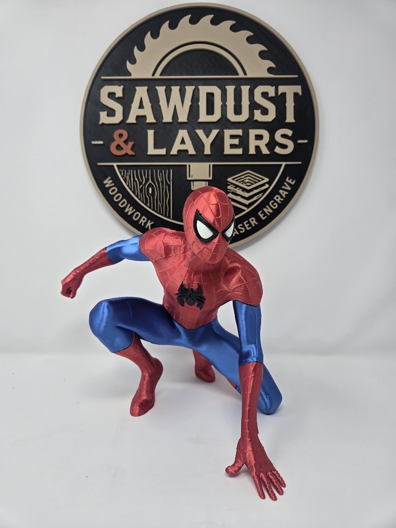 Puede incluir: Una figura de Spider-Man impresa en 3D en pose agachada con un traje rojo y azul met&aacute;lico y un emblema de ara&ntilde;a negro en el pecho. Detr&aacute;s de la figura hay un letrero circular negro y tostado con un gr&aacute;fico de sierra y el texto SAWDUST & LAYERS.