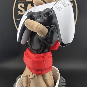 May include: A 3D printed figurine featuring a tan hand emerging from a rocky base to hold a white and black video game controller. The figure wears a black sleeve and a red belt, with a white circular border surrounding the base on a black background.