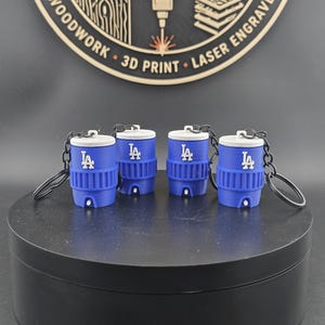 May include: Four blue and white miniature water cooler keychains featuring the LA logo on the front. Each keychain has a white lid, a blue ribbed body, a small white spigot at the base, and a black metal ring attached to the top for keys.