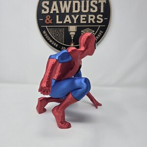 Puede incluir: Una figura impresa en 3D de un superh&eacute;roe en cuclillas, con un traje rojo y azul met&aacute;lico. La figura est&aacute; situada frente a un logotipo circular que dice SAWDUST & LAYERS, WOODWORK 3D GRAVE, con un gr&aacute;fico de una hoja de sierra en la parte superior.