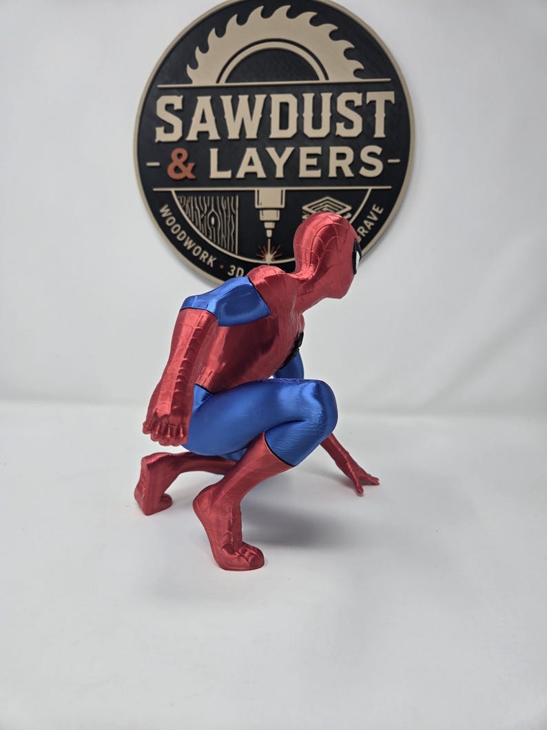 Puede incluir: Una figura impresa en 3D de un superh&eacute;roe en cuclillas, con un traje rojo y azul met&aacute;lico. La figura est&aacute; situada frente a un logotipo circular que dice SAWDUST & LAYERS, WOODWORK 3D GRAVE, con un gr&aacute;fico de una hoja de sierra en la parte superior.