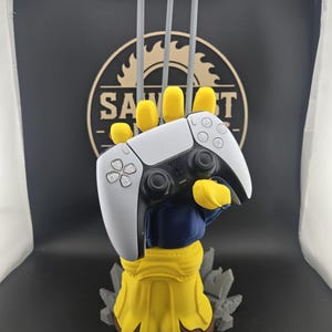 May include: A yellow and blue hand-shaped controller stand featuring three long grey claws extending upward. The stand holds a white and black wireless game controller. The base is yellow with a brown bottom and grey jagged rock-like accents.