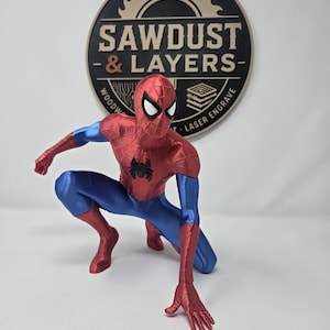Puede incluir: Una figura de Spider-Man en posici&oacute;n agachada con un traje rojo y azul met&aacute;lico, con un emblema de ara&ntilde;a negro en el pecho y ojos blancos. Detr&aacute;s de la figura hay un letrero circular negro y beige con el texto SAWDUST & LAYERS.