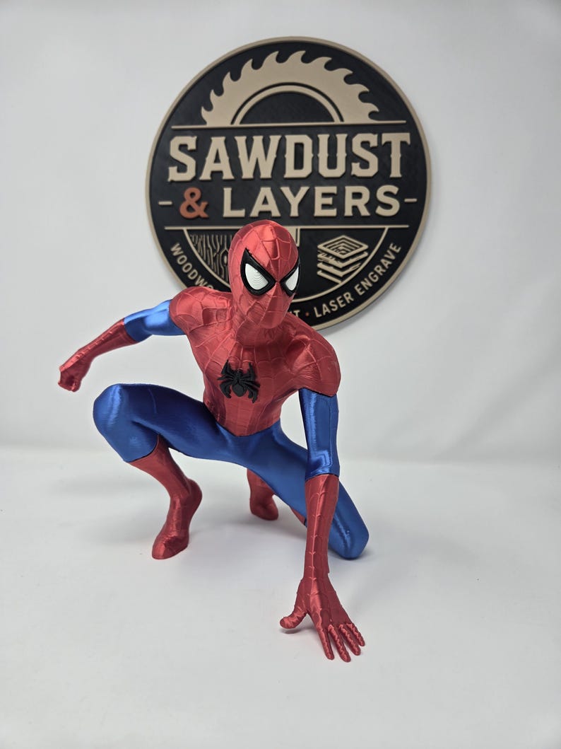 Puede incluir: Una figura de Spider-Man en posici&oacute;n agachada con un traje rojo y azul met&aacute;lico, con un emblema de ara&ntilde;a negro en el pecho y ojos blancos. Detr&aacute;s de la figura hay un letrero circular negro y beige con el texto SAWDUST & LAYERS.