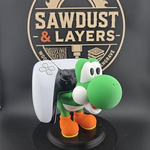 May include: A green and white Yoshi character figurine acting as a controller stand for a white and black video game controller. The figure stands on a circular black base in front of a circular logo featuring the text SAWDUST & LAYERS.