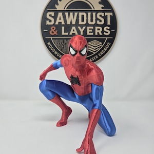 Puede incluir: Una figura de Spider-Man roja y azul en posici&oacute;n agachada con un emblema de ara&ntilde;a negro en el pecho. Detr&aacute;s de la figura hay un letrero circular negro y tostado con el texto SAWDUST & LAYERS WOODWORK LASER ENGRAVE en disposici&oacute;n circular.