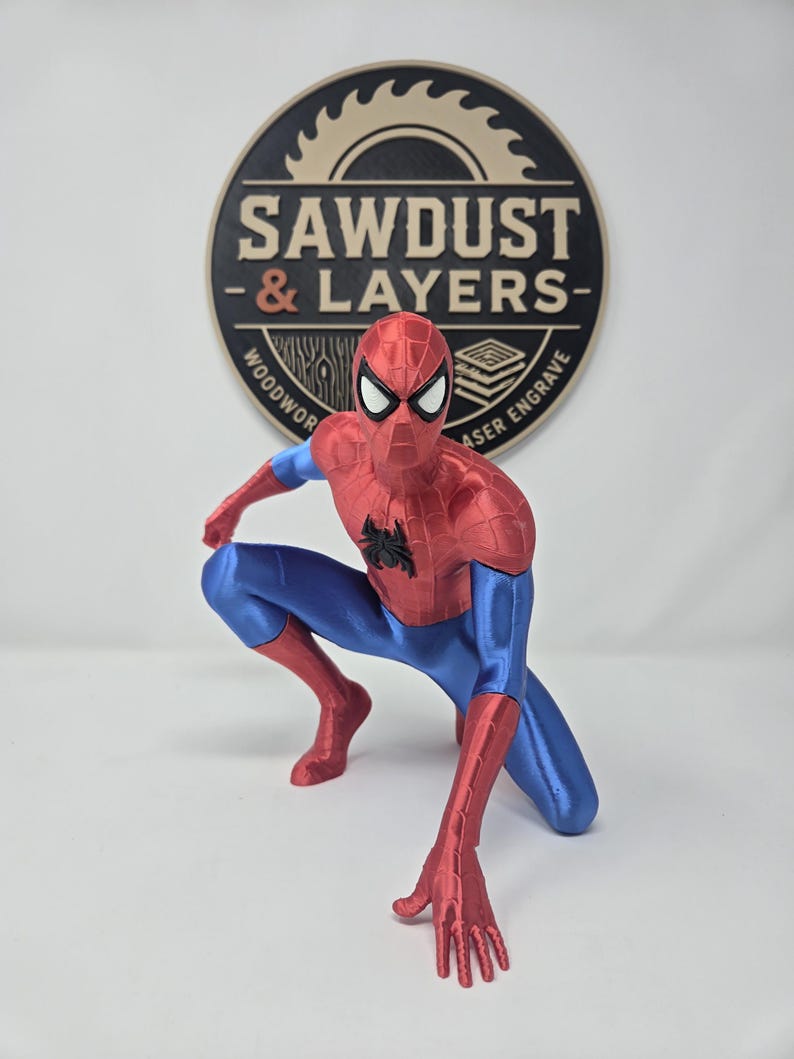 Puede incluir: Una figura de Spider-Man roja y azul en posici&oacute;n agachada con un emblema de ara&ntilde;a negro en el pecho. Detr&aacute;s de la figura hay un letrero circular negro y tostado con el texto SAWDUST & LAYERS WOODWORK LASER ENGRAVE en disposici&oacute;n circular.