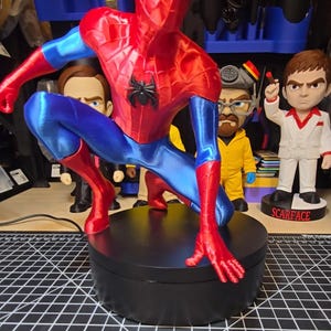 Puede incluir: Una figura de Spider-Man impresa en 3D y brillante en pose agachada, con un traje rojo vibrante y azul met&aacute;lico con un emblema de ara&ntilde;a negro en el pecho. La figura est&aacute; montada sobre una base de exhibici&oacute;n circular de color negro.