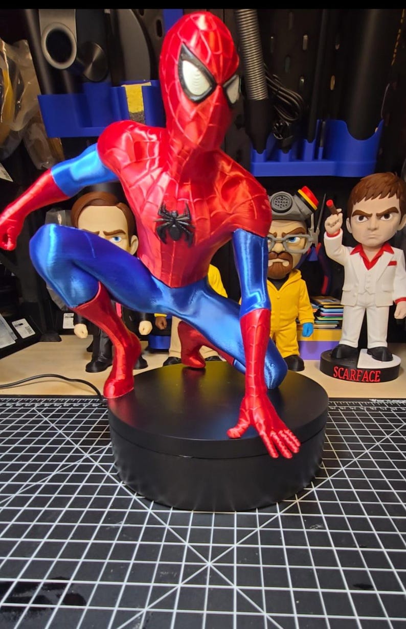 Puede incluir: Una figura de Spider-Man impresa en 3D y brillante en pose agachada, con un traje rojo vibrante y azul met&aacute;lico con un emblema de ara&ntilde;a negro en el pecho. La figura est&aacute; montada sobre una base de exhibici&oacute;n circular de color negro.