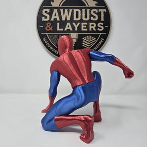 Puede incluir: Una figura impresa en 3D de un superh&eacute;roe en posici&oacute;n agachada, con un traje met&aacute;lico rojo y azul. La figura est&aacute; colocada frente a un logotipo circular que dice SAWDUST & LAYERS WOODWORK ENGRAVE con un gr&aacute;fico de hoja de sierra.