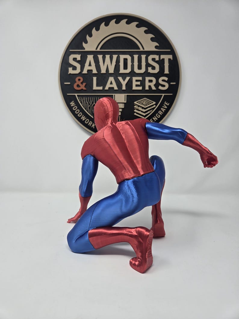 Puede incluir: Una figura impresa en 3D de un superh&eacute;roe en posici&oacute;n agachada, con un traje met&aacute;lico rojo y azul. La figura est&aacute; colocada frente a un logotipo circular que dice SAWDUST & LAYERS WOODWORK ENGRAVE con un gr&aacute;fico de hoja de sierra.