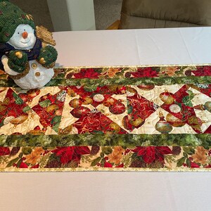 May include: A quilted table runner with a Christmas theme. The runner features a cream background with red, green, and gold ornaments. The runner is trimmed with a green, red, and gold border.