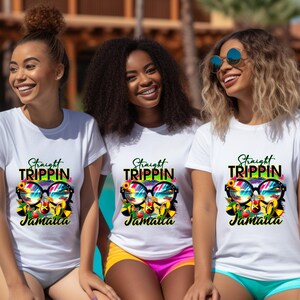 May include: Three women wearing white t-shirts with a colorful graphic design. The design includes sunglasses, a beach scene, and the words "Straight Trippin' Jamaica". The t-shirts are short-sleeved.