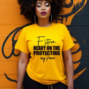 May include: A yellow t-shirt with the text "Extra Heavy On The Protecting my Peace" in black script. The model is wearing black pants and large silver hoop earrings. The background is an orange and black mural.